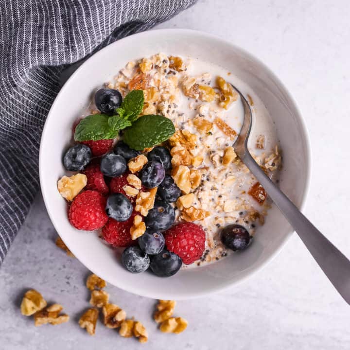 White bowl of homemade muesli with fresh berries, mint, walnuts, and a silver spoon and blue striped linen