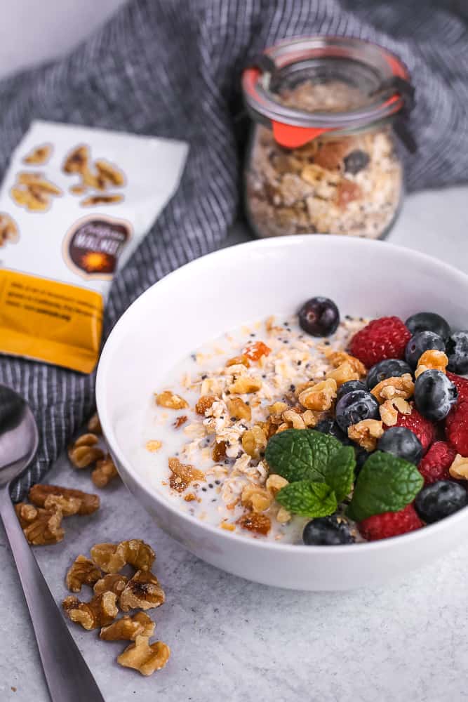 White bowl of homemade muesli with a spoon, extra walnuts, and package of extra muesli ingredients on a light grey background