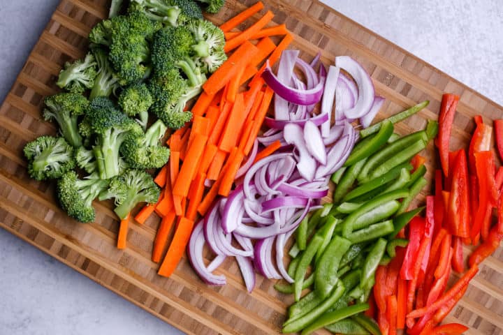 Colorful veggies in a butcher block cutting board, including broccoli, carrots, onions, and red and green bell peppers