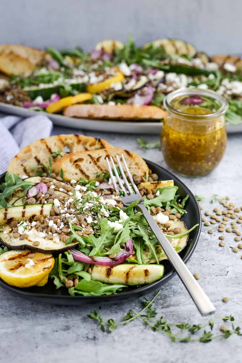 A black ceramic salad plate is placed on a kitchen countertop, filled with a vegetarian summer salad with brown lentils, grilled vegetables with prominent char marks, feta cheese, and a light dressing. Grilled baguette slices are arranged on the side of the plate with a silver fork