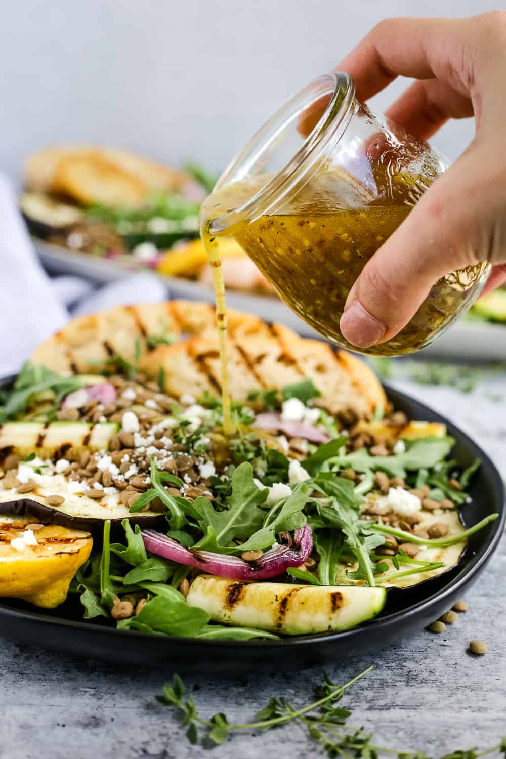 A woman's hand holds a small glass jar full of a honey mustard vinaigrette salad dressing, and lightly pours it over a prepared salad on a black ceramic salad plate