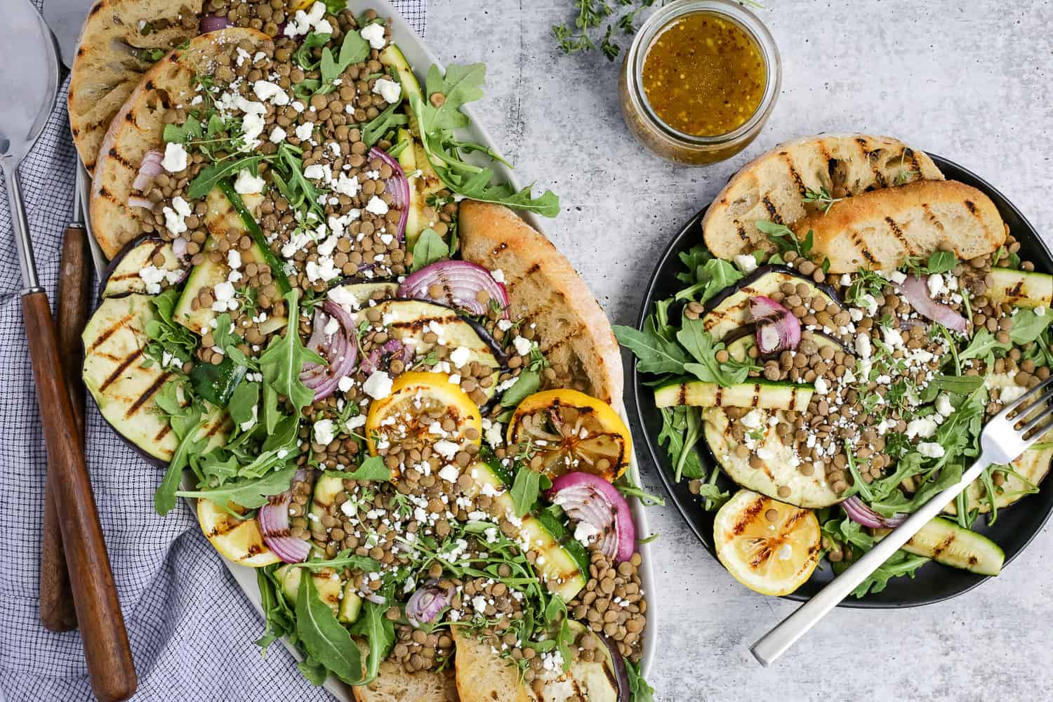 Horizontal overhead view of a green salad with lentils recipe, featuring grilled vegetables and feta cheese, displayed on a kitchen countertop with salad tongs and extra dressing nearby