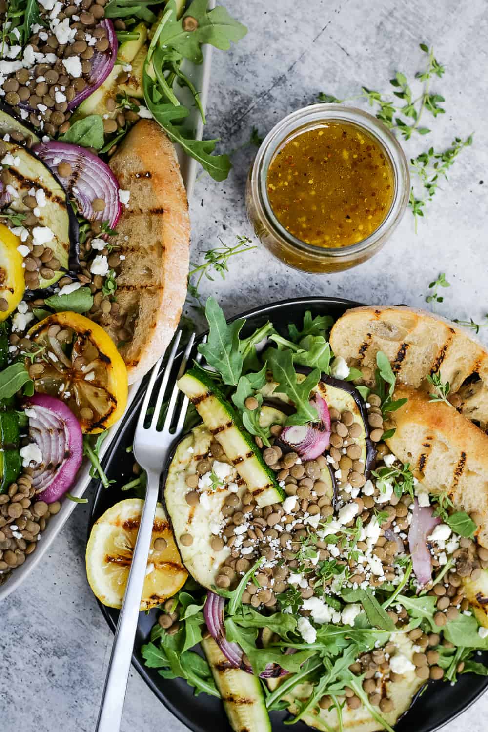 Overhead view of a serving platter and salad plate filled with a green salad with lentils, grilled vegetables, and feta cheese with a glass jar of dressing placed nearby on a kitchen countertop