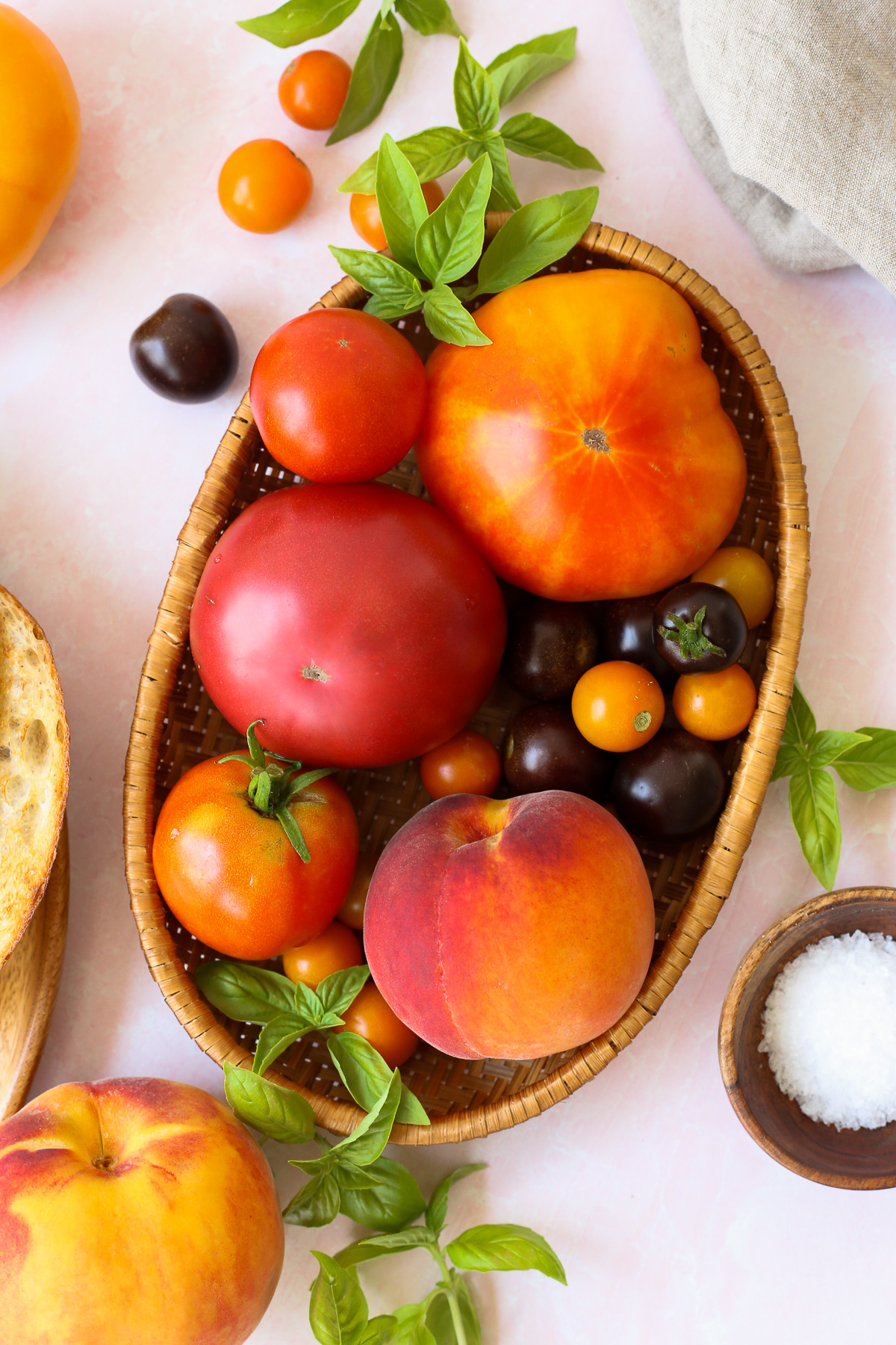 An assortment of ripe tomatoes and peaches in varying shades of orange, displayed in a woven basket with sprigs of fresh basil scattered nearby.