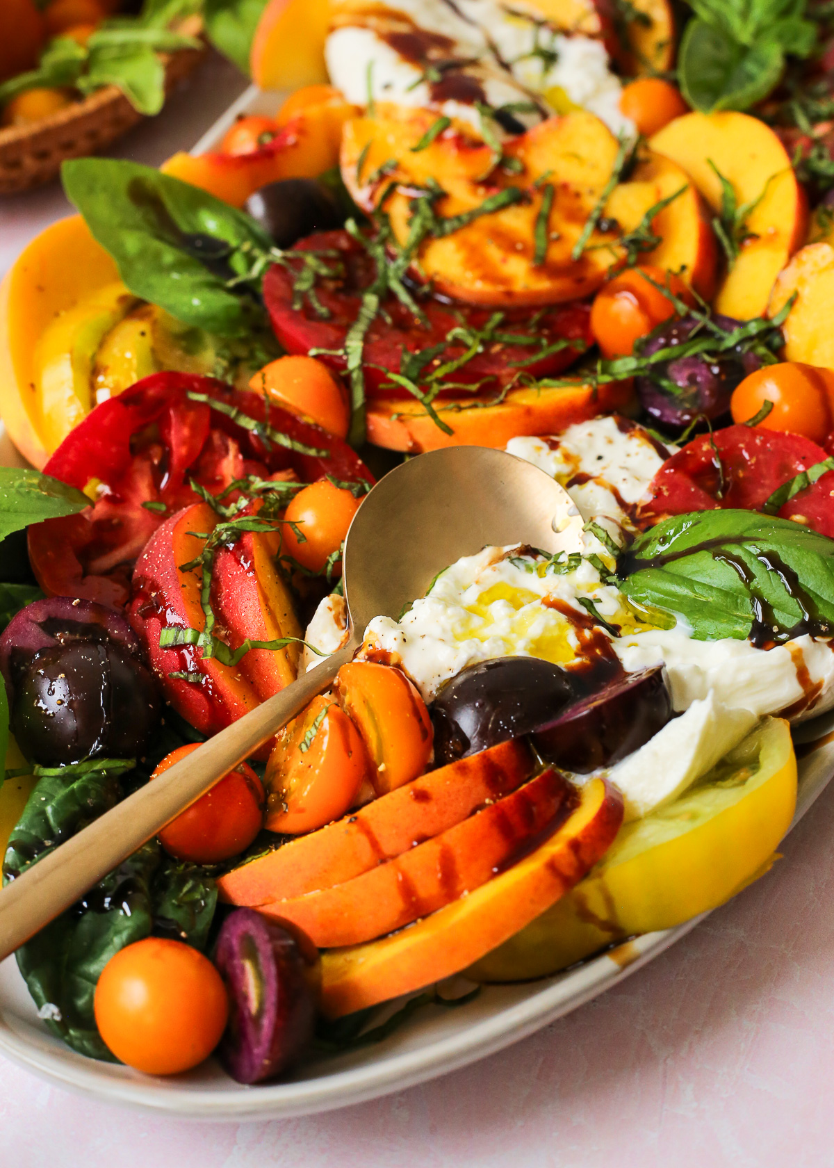 A large serving spoon breaks open a ball of burrata cheese that's sitting between slices of fresh peaches and tomatoes on a large ceramic serving platter.