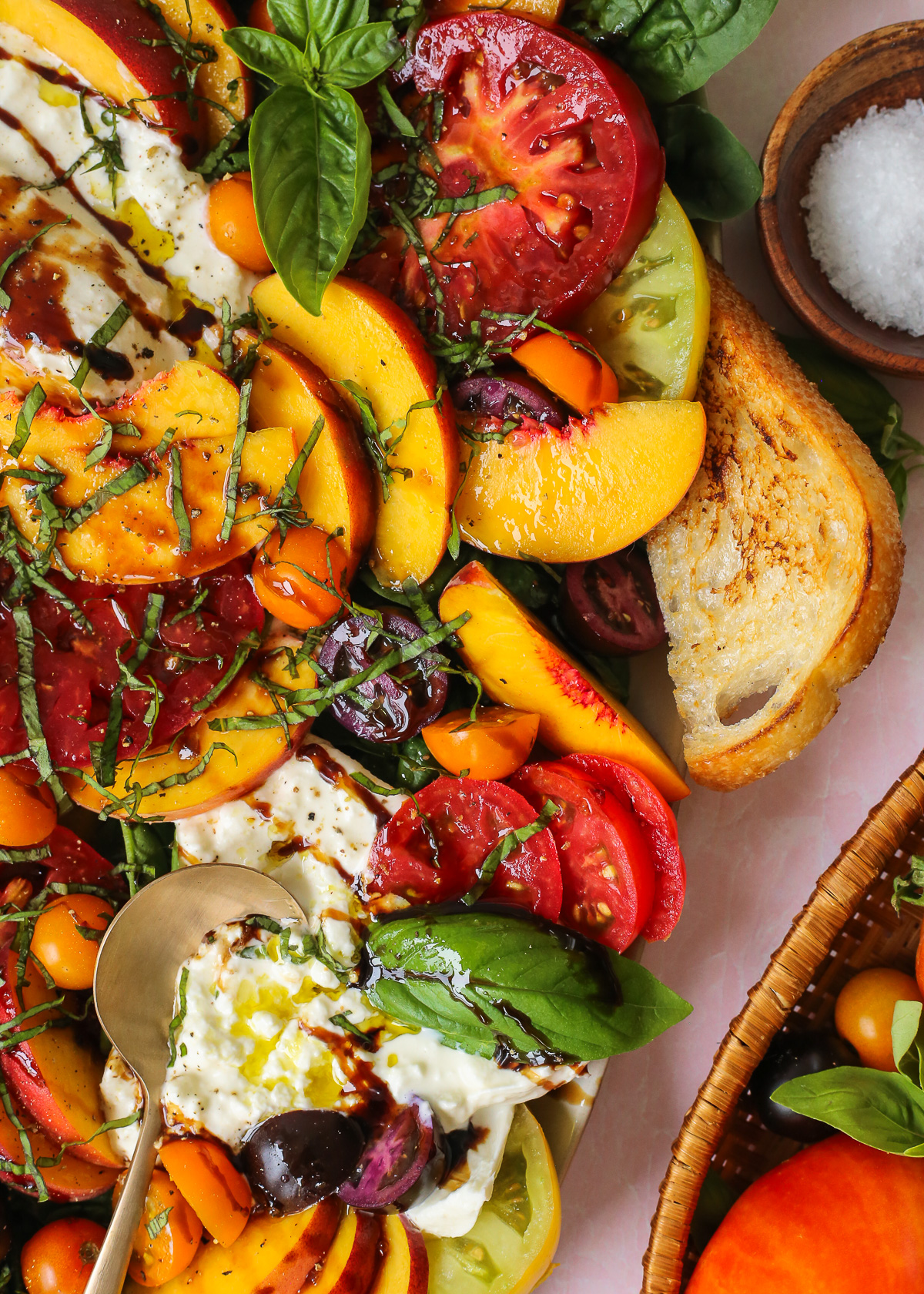 A large serving spoon rests inside a ball of burrata, nestled between large slices of fresh peaches and tomatoes and garnished with basil and balsamic glaze.