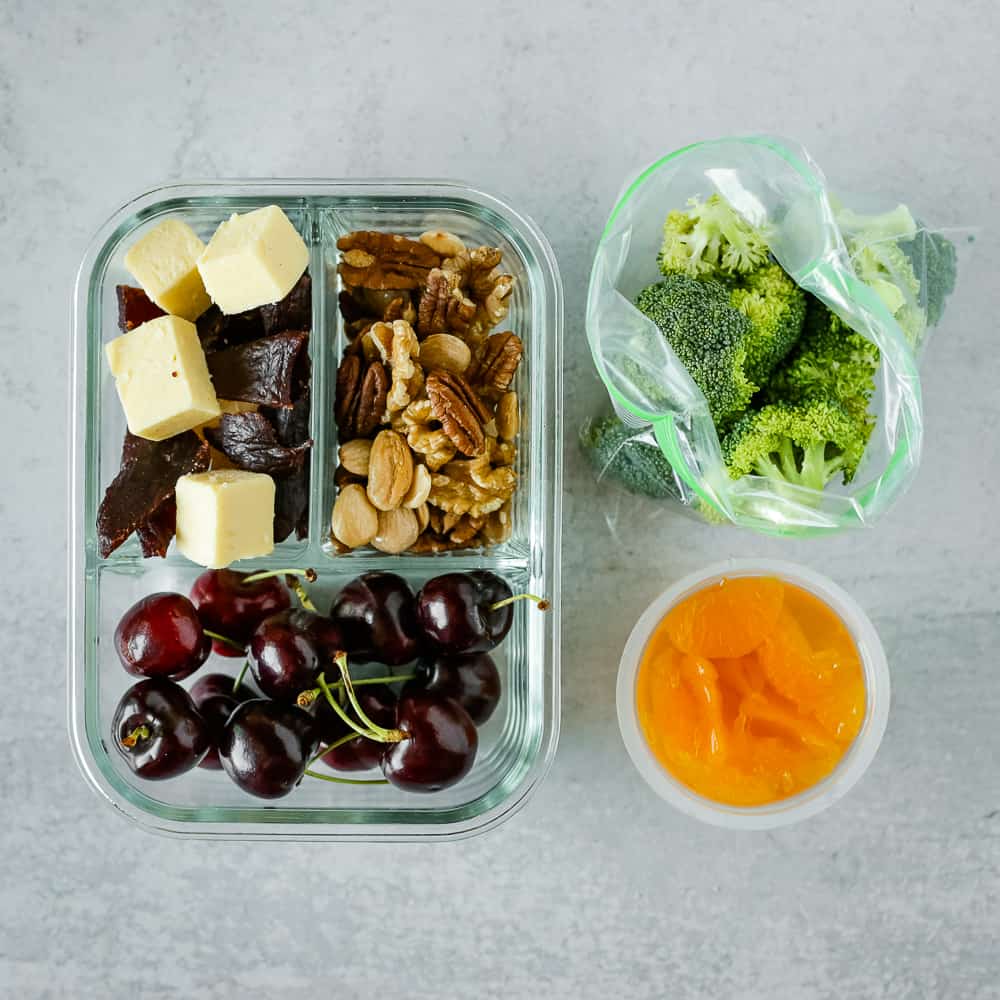 Snack lunch prepped in a glass storage container with fresh cherries, beef jerky and cheese cubes, mixed nuts, mandarin oranges, and broccoli