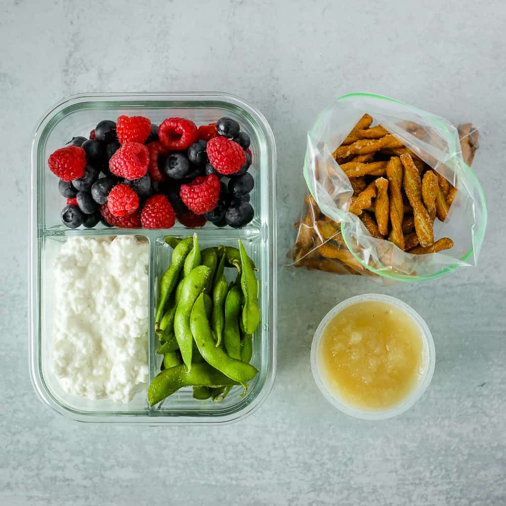 Snack meal prepped in a glass storage container with raspberries, blueberries, cottage cheese, edamame, pretzels, and applesauce