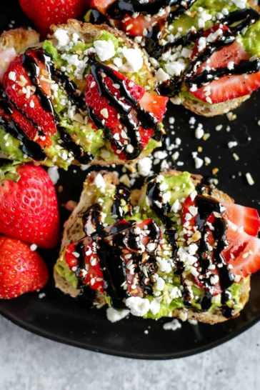 An overhead view of a black plate with avocado toast topped with sliced strawberries, feta cheese, and balsamic glaze