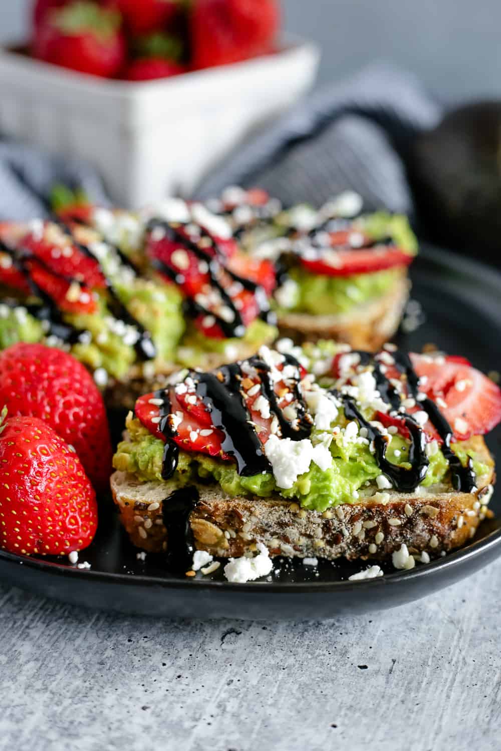 A black plate with two thick slices of avocado toast topped with sliced strawberries, feta cheese, and balsamic glaze