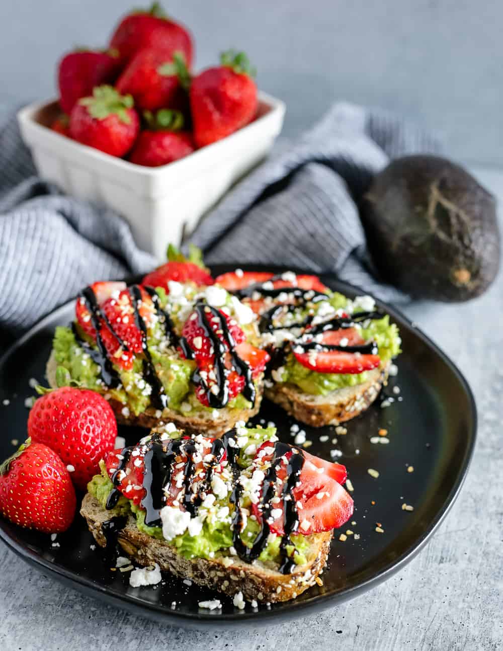 Three thick slices of avocado toast on a black plate, topped with sliced strawberries, feta cheese, and balsamic glaze