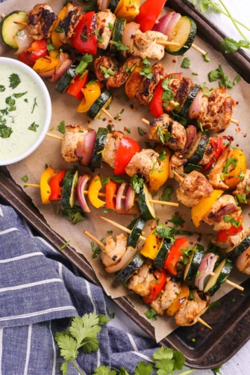 Yogurt marinated chicken kebabs on a serving platter with grilled veggies and creamy dipping sauce, overhead shot