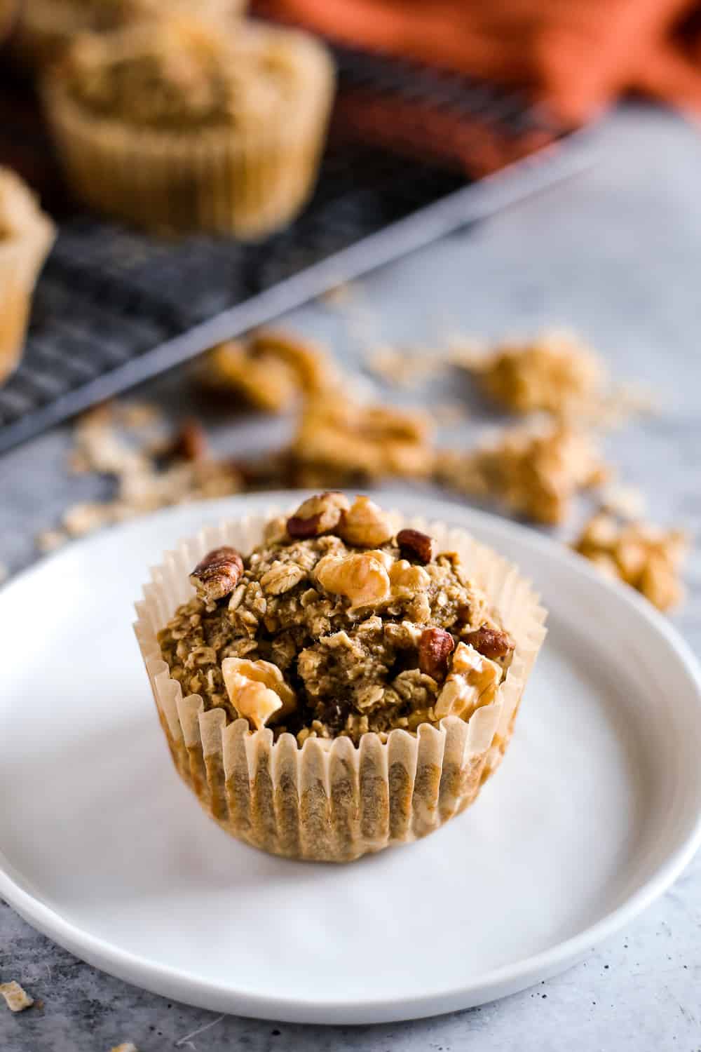 A banana nut baked oatmeal cup in a cupcake liner sits on a small white dish in front of a cooling rack with the rest of the muffins