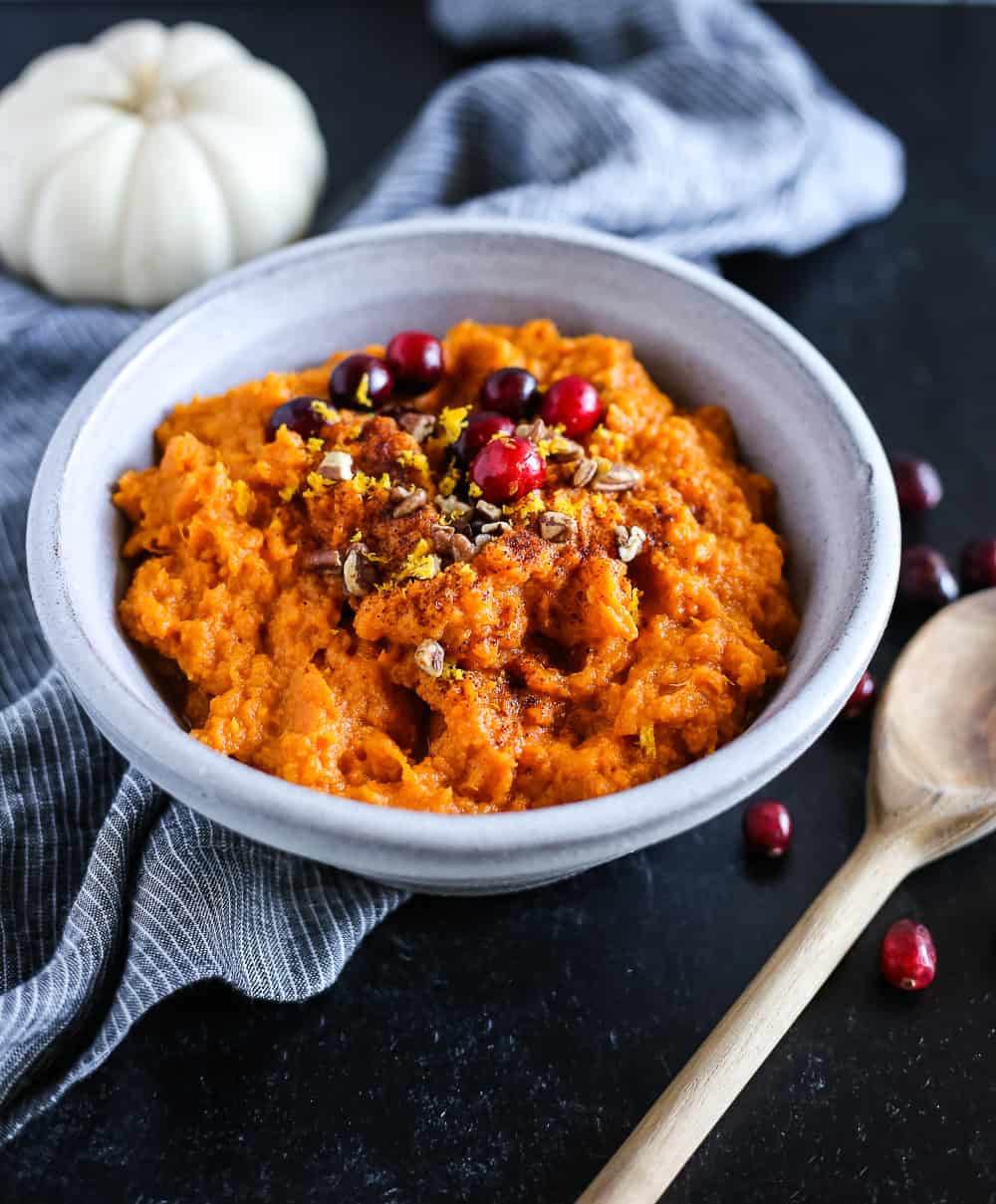 Mashed sweet potatoes in a pale blue ceramic serving bowl, garnished with chopped pecans and fresh cranberries on a black countertop