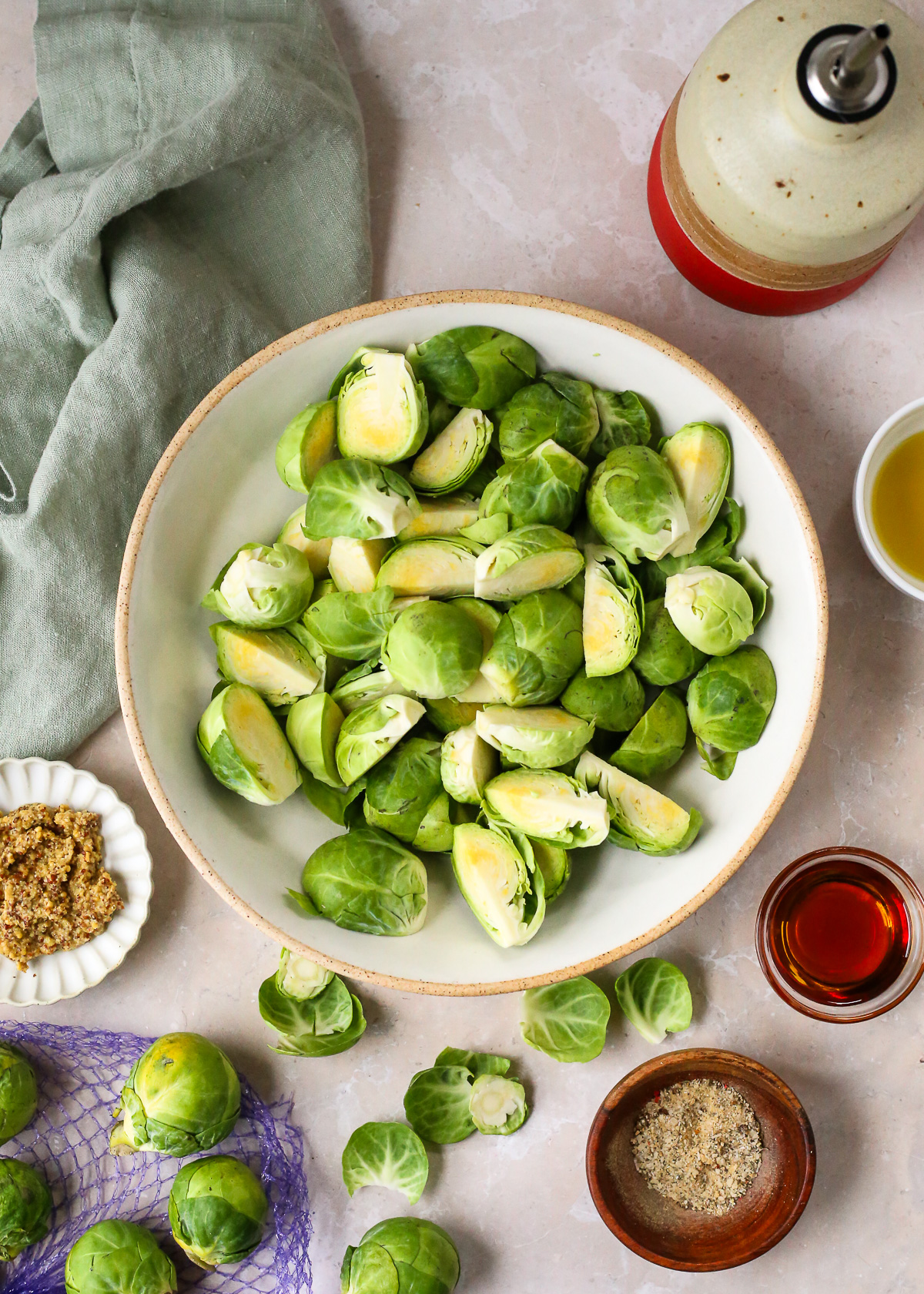A large prep bowl filled with uncooked Brussels sprouts, trimmed and sliced, on a kitchen countertop with various small prep bowls placed nearby. 
