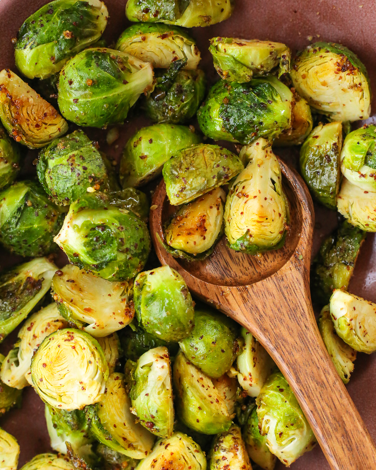 A wooden serving spoon rests in a bowl filled with roasted Brussels sprouts coated in a maple mustard sauce. 
