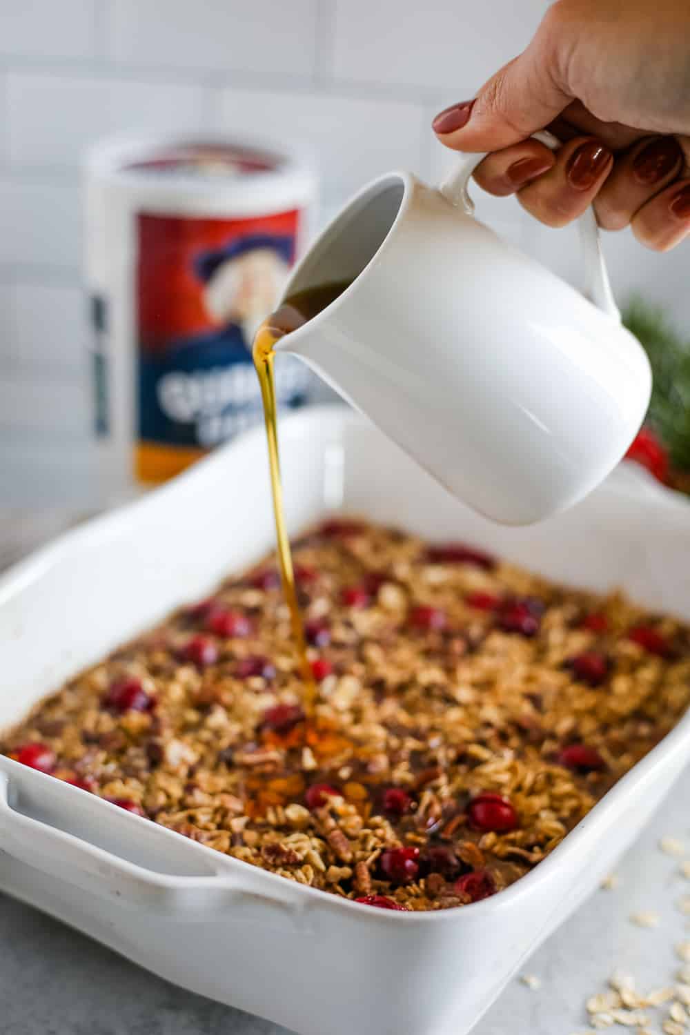 A woman's hand pours maple syrup over a baked oatmeal recipe with a canister of Quaker Oats in the background