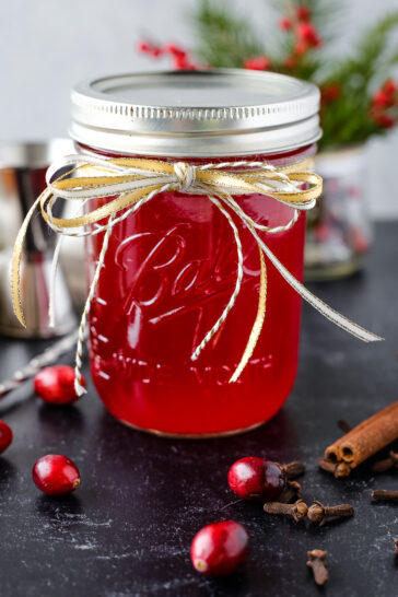 A glass mason jar with a bright red cranberry simple syrup, garnished with a gold ribbon on a black countertop
