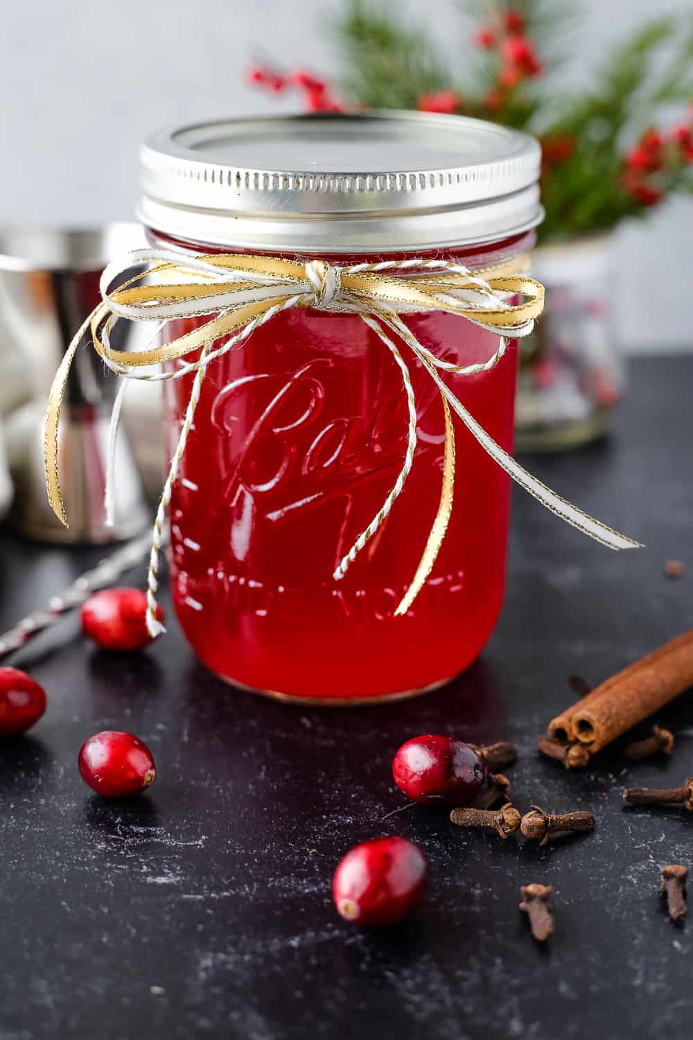 A glass mason jar with a bright red cranberry simple syrup, garnished with a gold ribbon on a black countertop