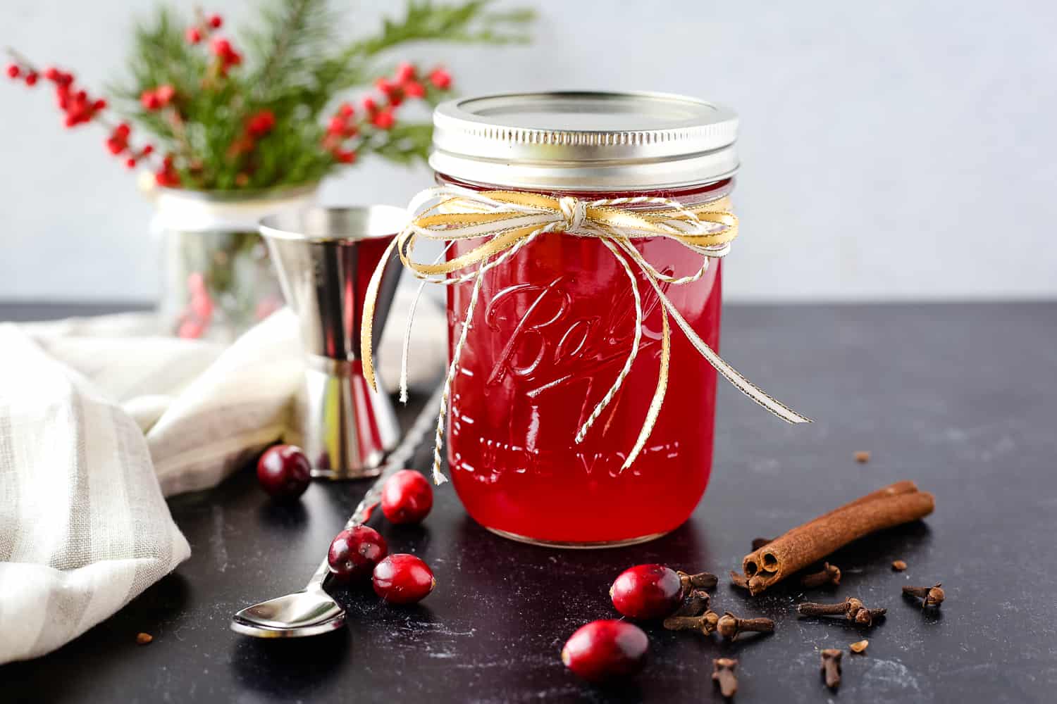 A glass mason jar filled to the top with a vibrant red spiced cranberry simple syrup is decorated with seasonal ribbon and displayed with a silver bar spoon and jigger