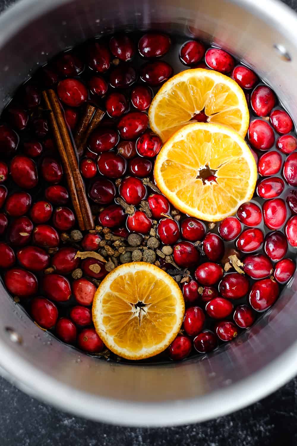 An overhead view of fresh cranberries, cinnamon sticks, and orange slices in a stainless steel cooking pot