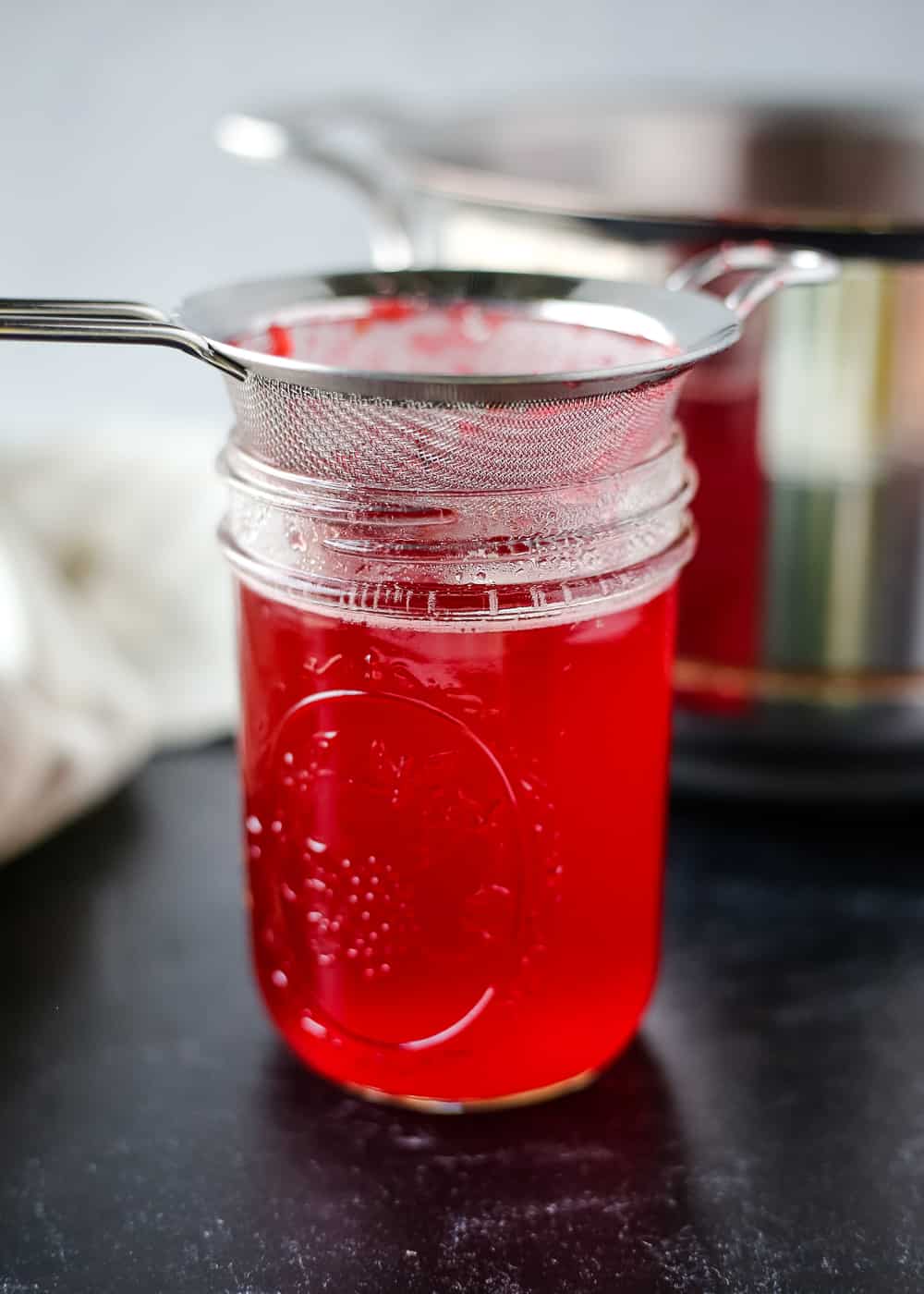 A glass mason jar full of a clear red liquid with a silver strainer sitting on the lip of the jar