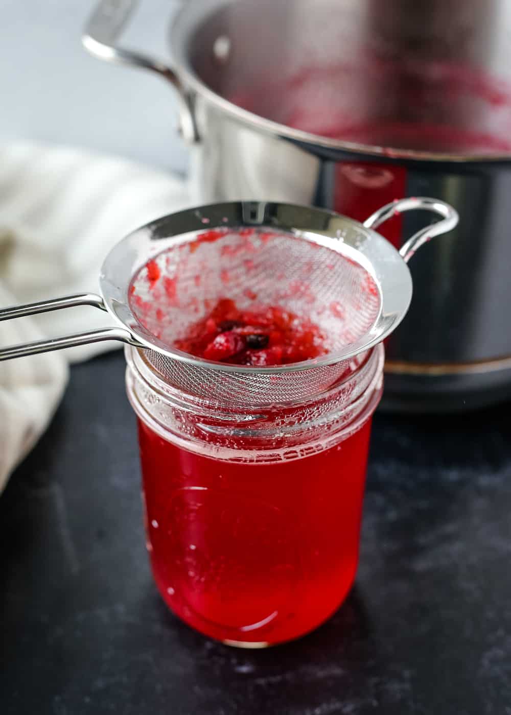 A glass mason jar full of cranberry simple syrup with a silver strainer and saucepan on a black countertop