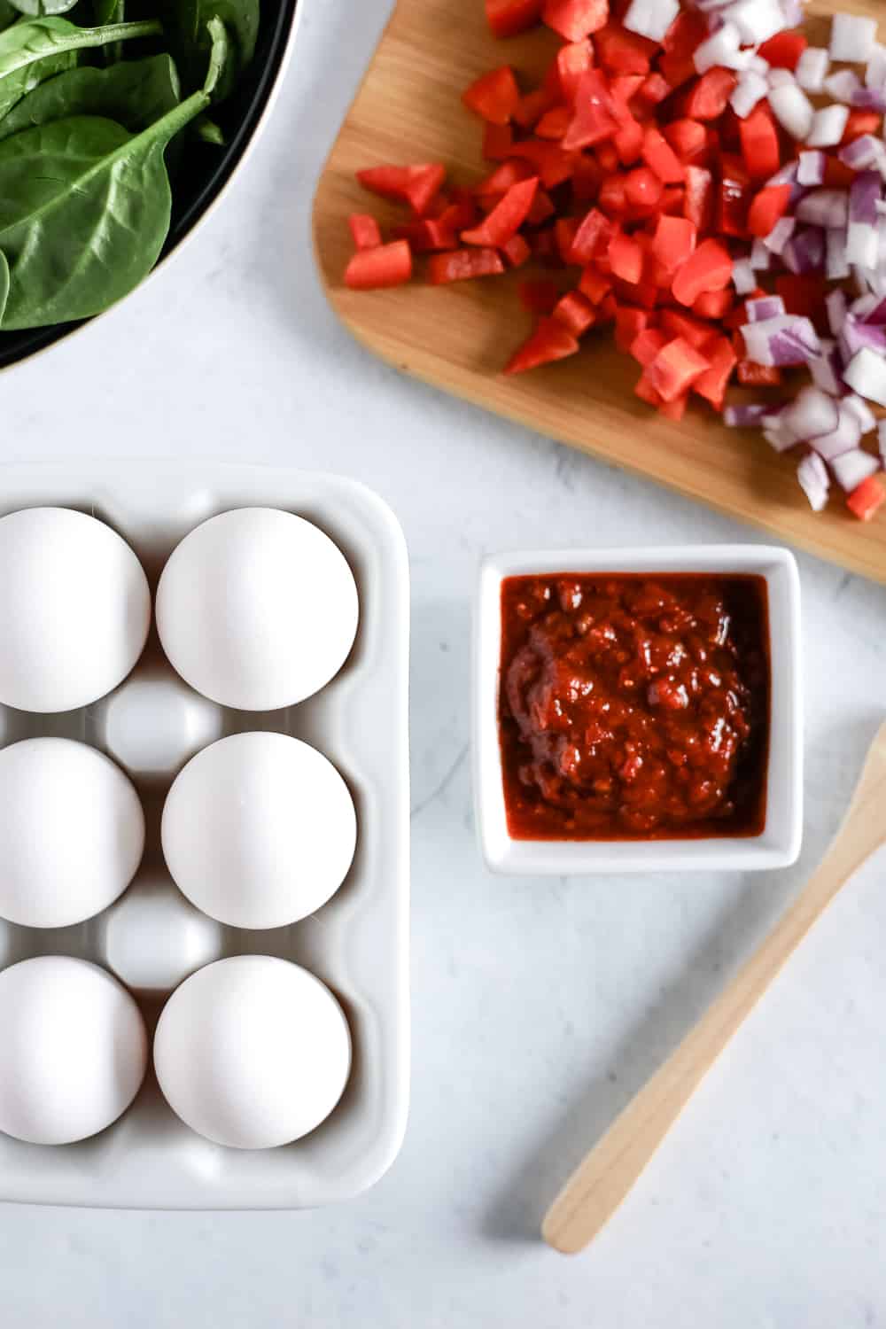 An overhead view of white eggs, fresh veggies, a cutting board, and a sauce ready to be cooked into a breakfast recipe