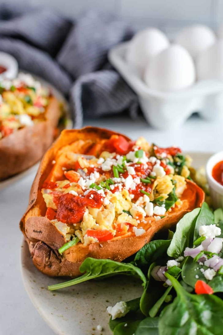 A stuffed sweet potato with scrambled eggs, spinach, and veggies on a plate on a kitchen countertop. A second serving is plated in the background