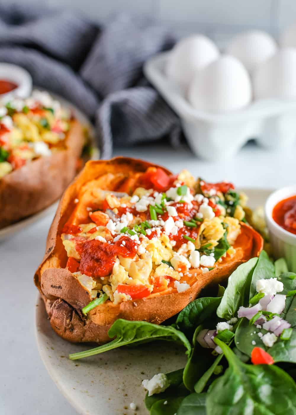 A stuffed sweet potato with scrambled eggs, spinach, and veggies on a plate on a kitchen countertop. A second serving is plated in the background