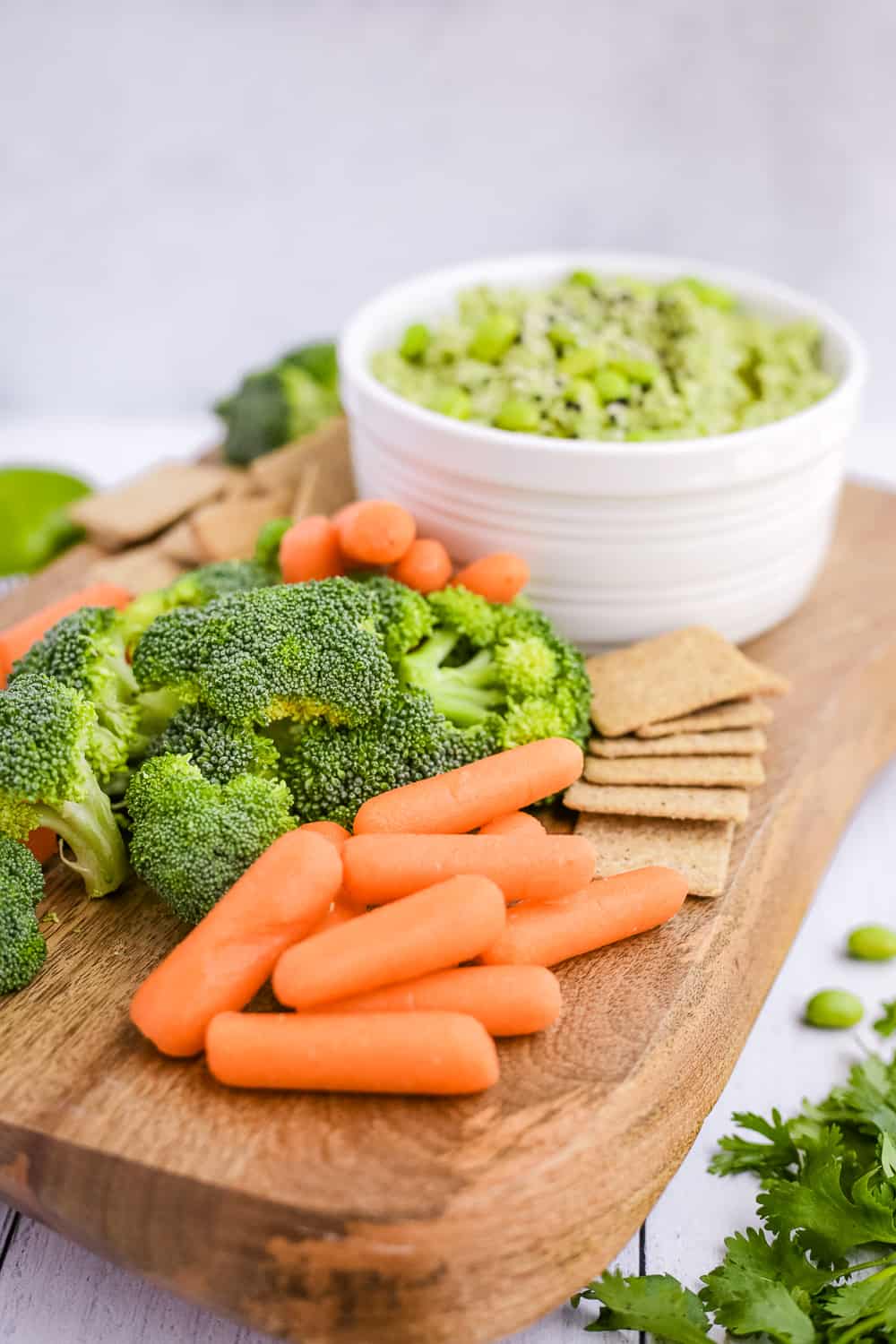 A wooden serving platter with carrots, broccoli, crackers, and a white bowl with hummus