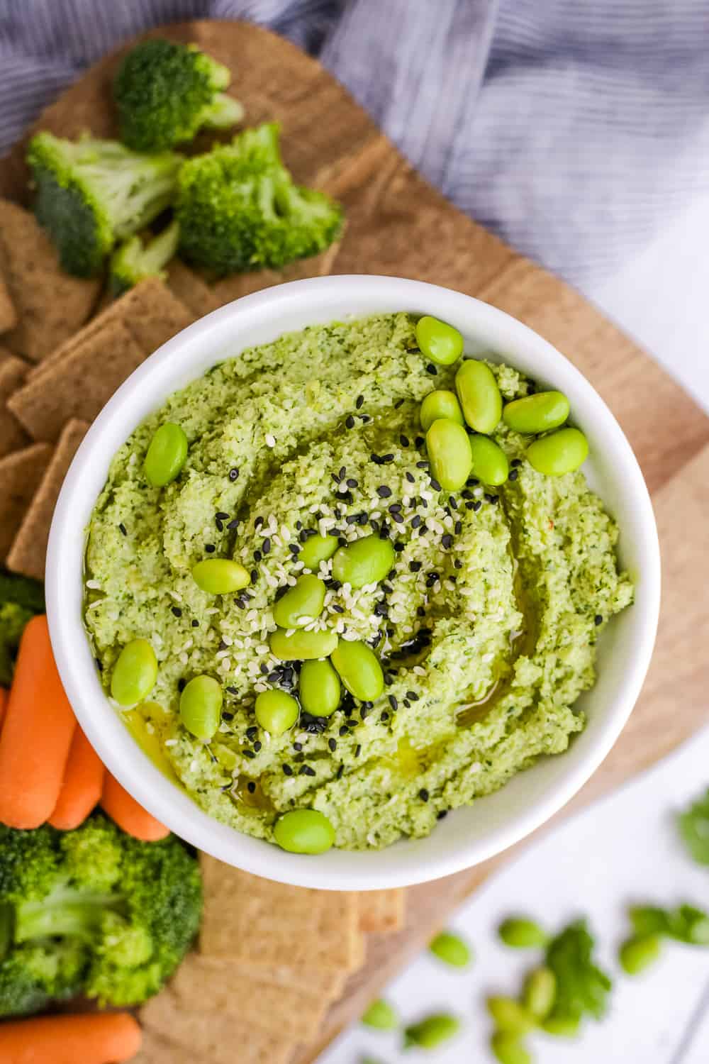 Overhead view of a bowl of edamame hummus on a serving board with fresh veggies and crackers