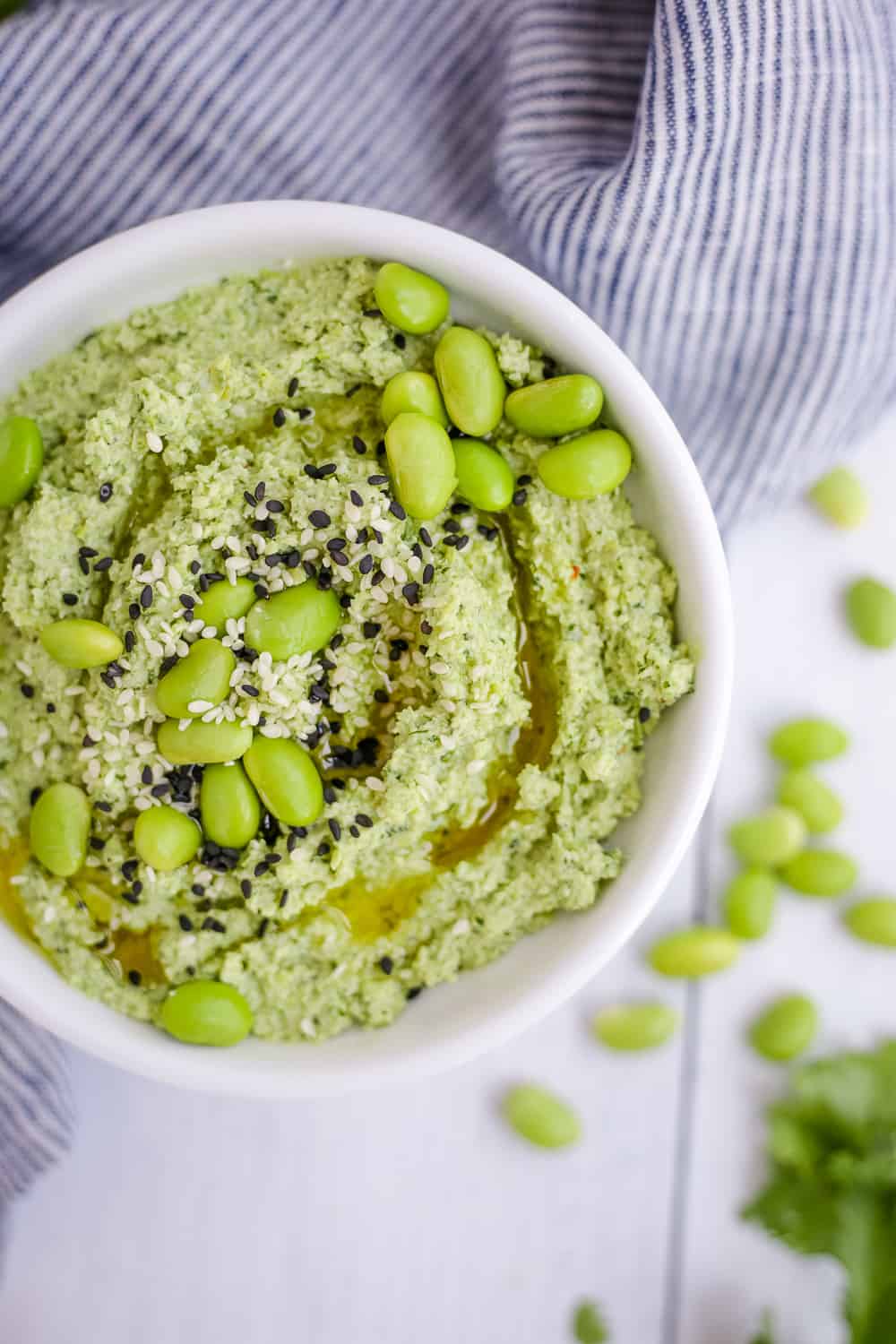 A white bowl of edamame hummus, overhead view, with shelled edamame and cilantro scattered around the bowl