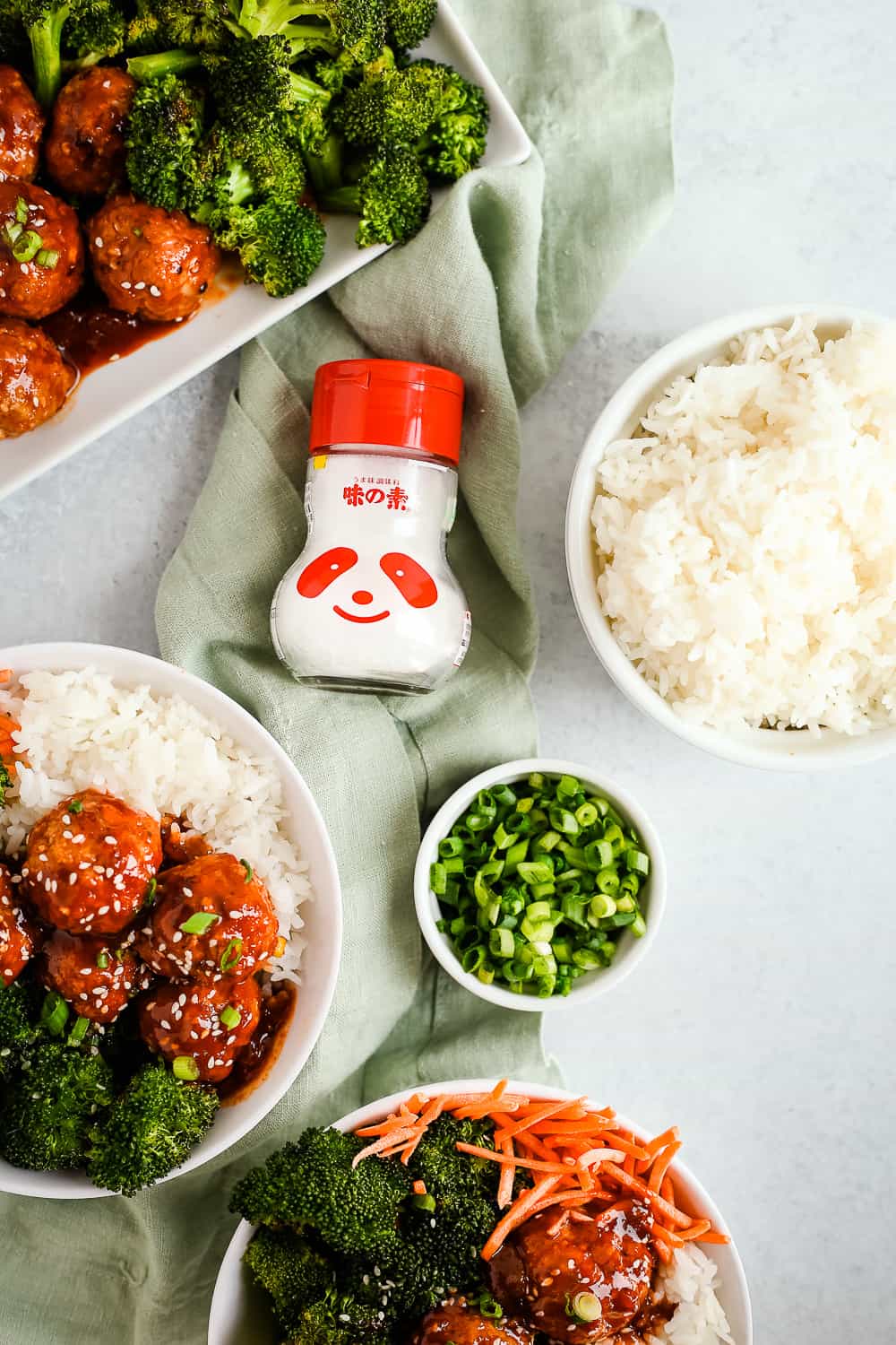 An overhead view of glass shaker of MSG with a red lid on a green linen in between dishes of rice bowls with spicy turkey meatballs and roasted broccoli