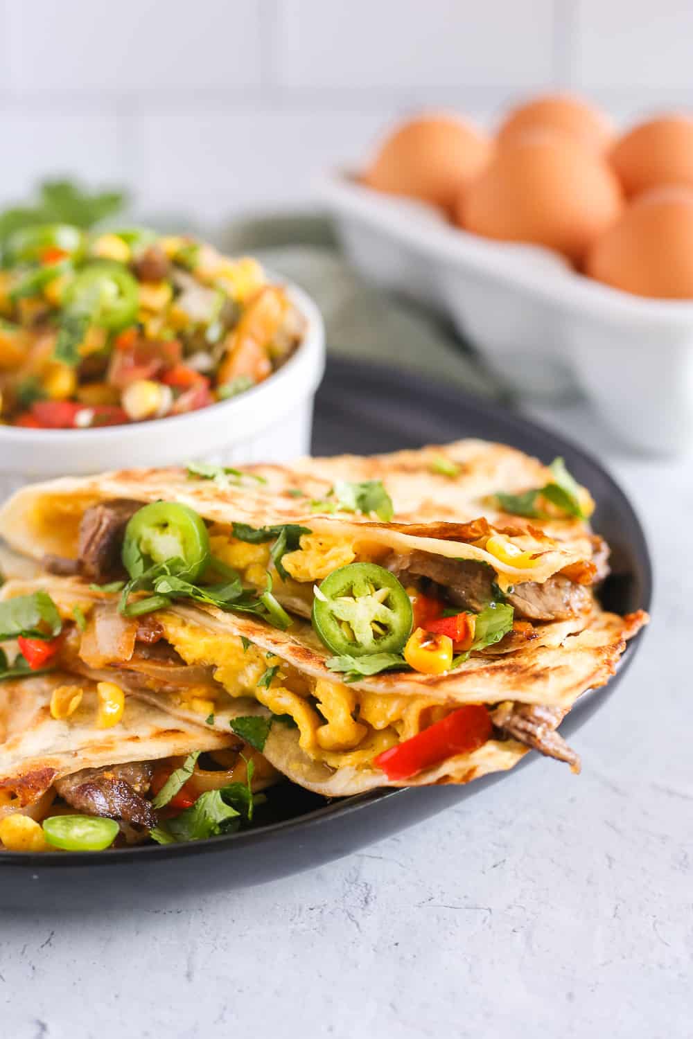 A close up shot of a breakfast beef quesadilla on a black plate. It is filled with colorful veggies and sliced hot peppers