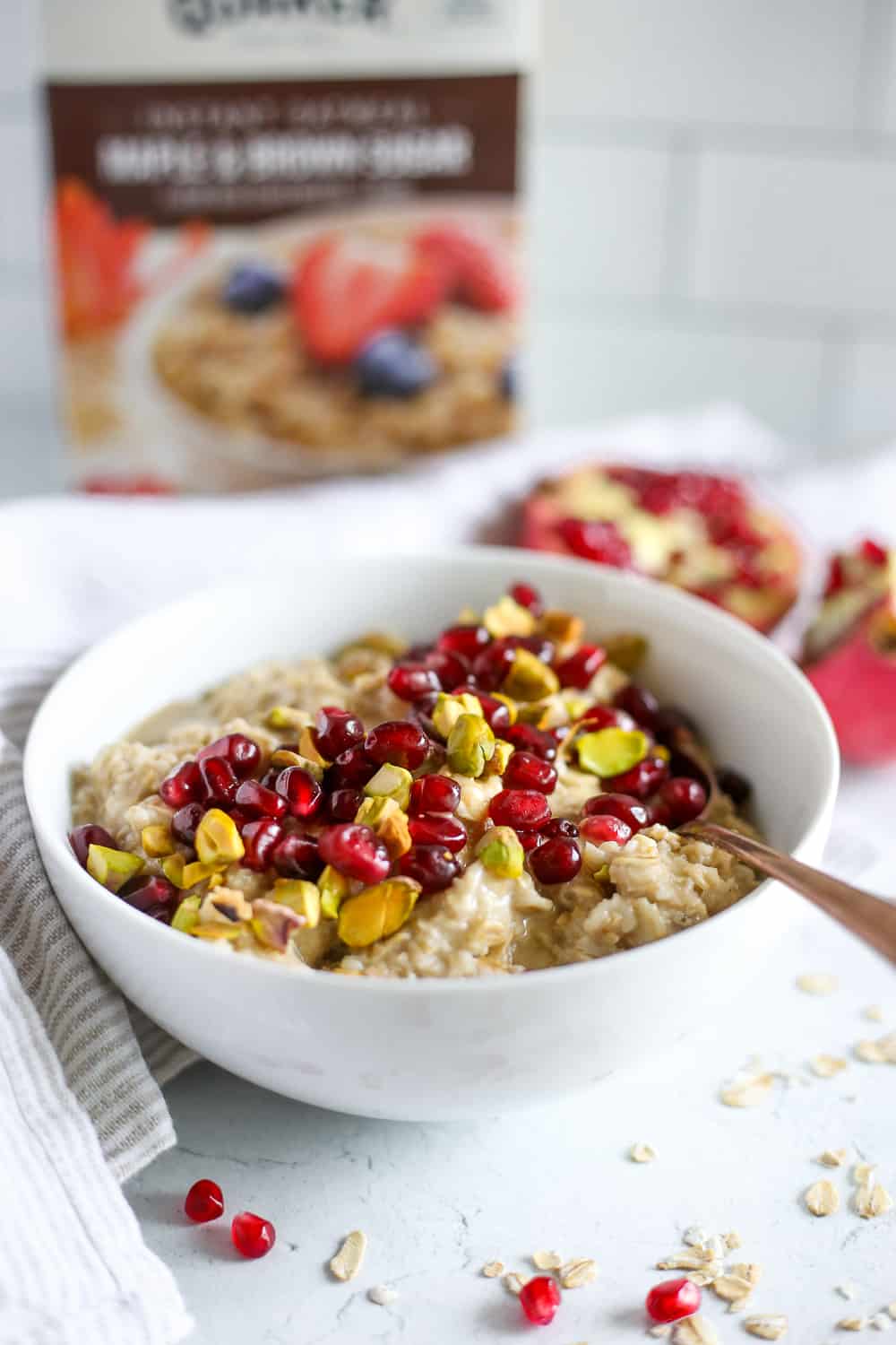 A view of a white bowl on a kitchen countertop, filled with a colorful hot oatmeal mixture topped with pomegranate, pistachios, and tahini with a box of Quaker oatmeal in the background