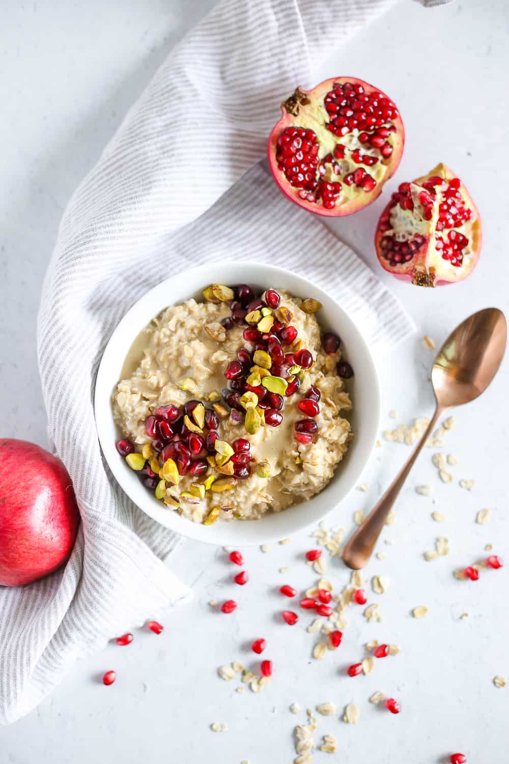 An overhead view of a white bowl on a kitchen countertop, filled with Maple-Tahini Oatmeal with pomegranates and chopped pistachios, surrounded by a partial pomegranate and a copper colored spoon