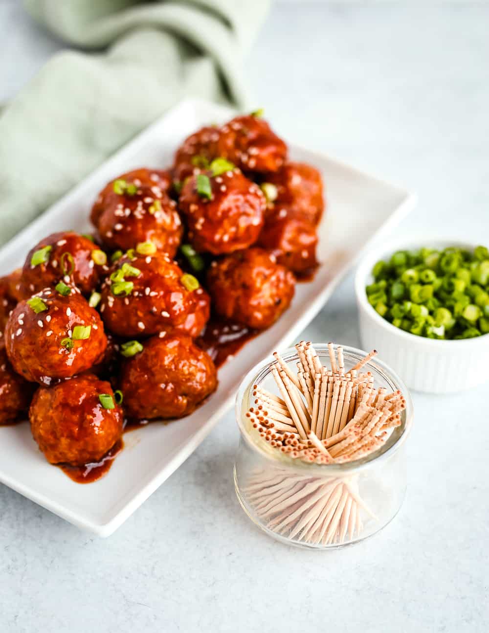 Photo of glazed spicy turkey meatballs on a white serving platter with green onions for garnish and a small bowl of toothpicks