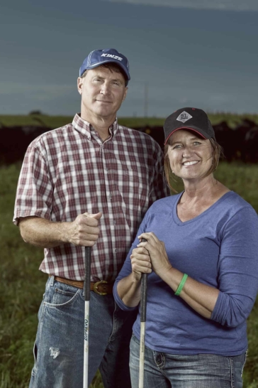 Two Kansas beef ranchers, a man and a woman, stand in a field and face the camera. They are smiling and wearing work clothing while holding tools