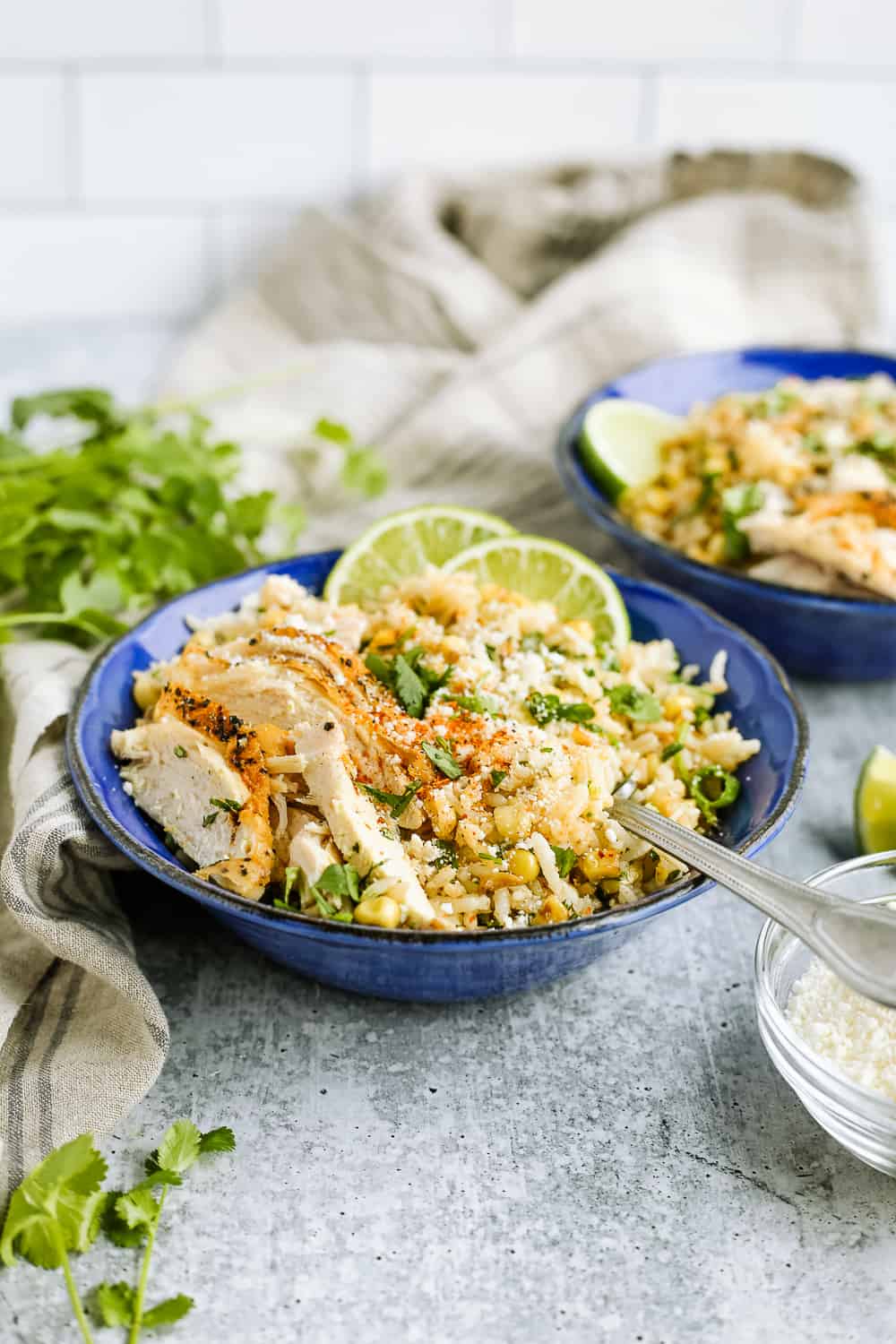 A blue bowl filled with rice, corn, sliced chicken, and lime wedges on a kitchen countertop with extra cilantro in the background