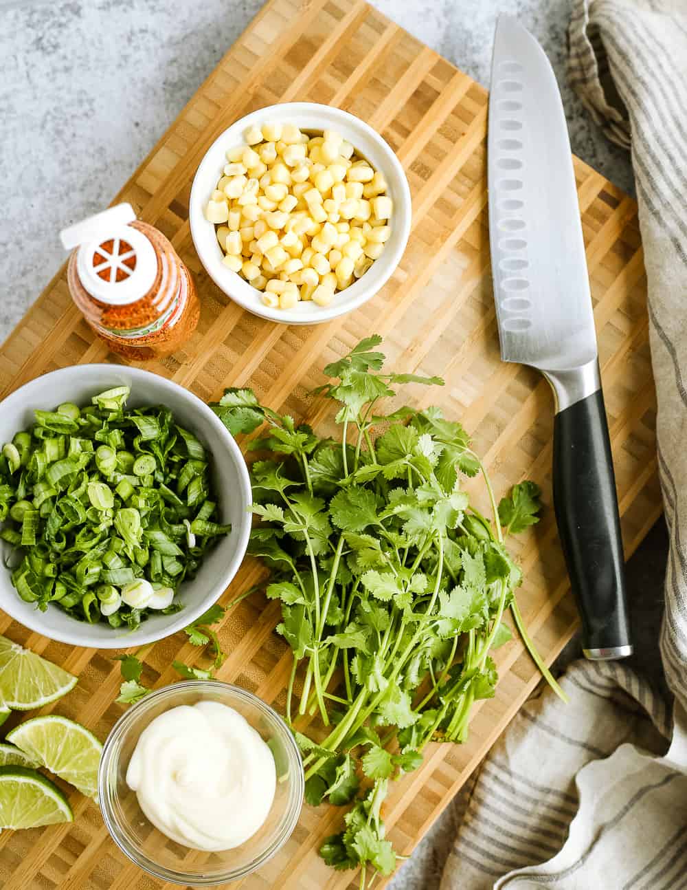 Overhead view of a butcher block cutting board with chopped green onions, fresh cilantro, sweet corn, lime wedges, a small clear glass bowl of mayo, and a sharpened chefs knife