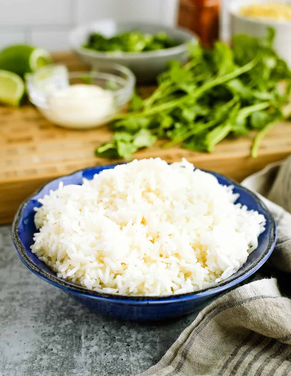 Close crop view of cooked long-grain white rice in a blue ceramic bowl in front of a butcher block cutting board with esquites-inspired ingredients and cilantro