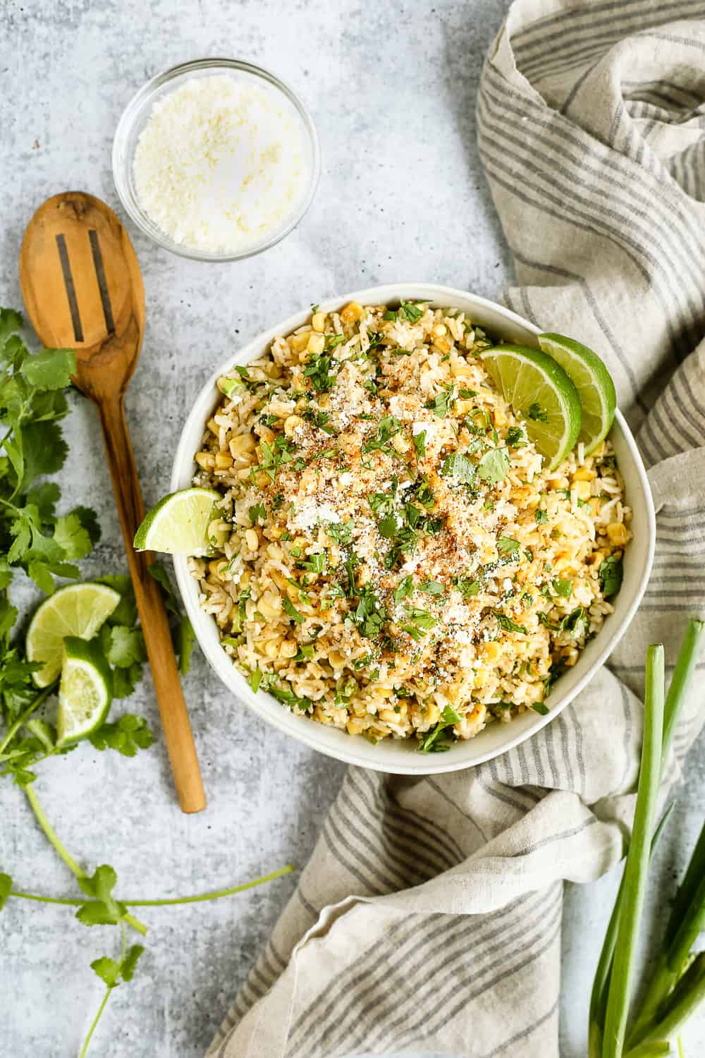 Overhead view of esquites-inspired Mexican street corn and rice bowls, with a wooden serving spoon, grey striped linen, and fresh herbs surrounding the serving bowl