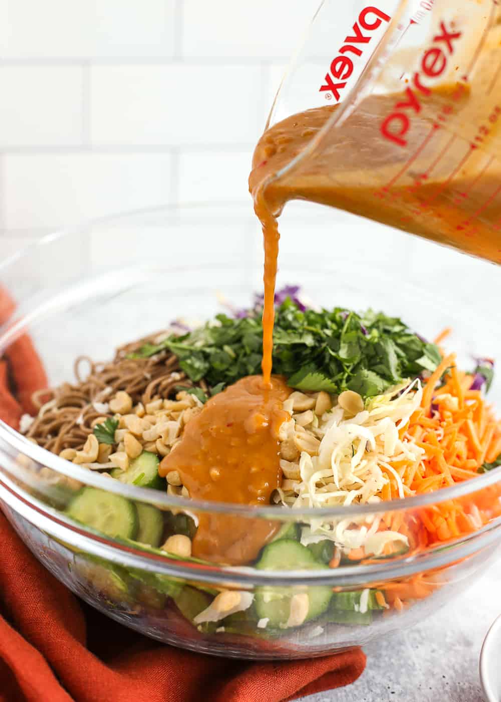 Creamy peanut sauce is poured from a clear glass measuring cup into a mixing bowl with soba noodles, sliced cucumbers, chopped cilantro, and shredded cabbage and carrots