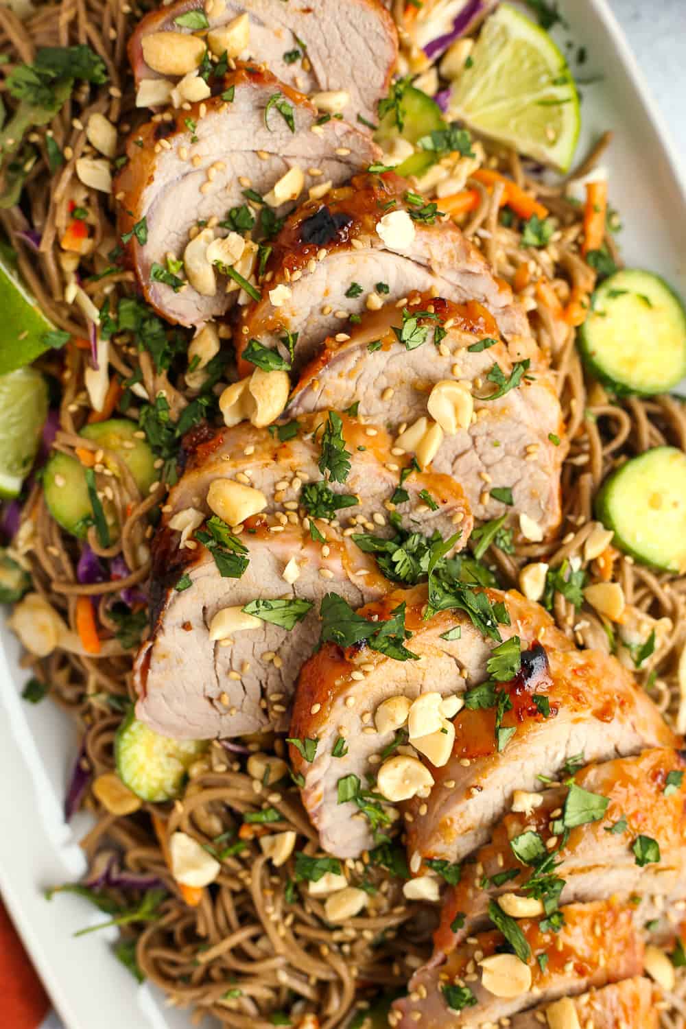 A closeup, overhead view of Soba Noodle Salad with Peanut Sauce and Pork, plated on a serving dish and garnished with chopped peanuts, sesame seeds, and lime wedges