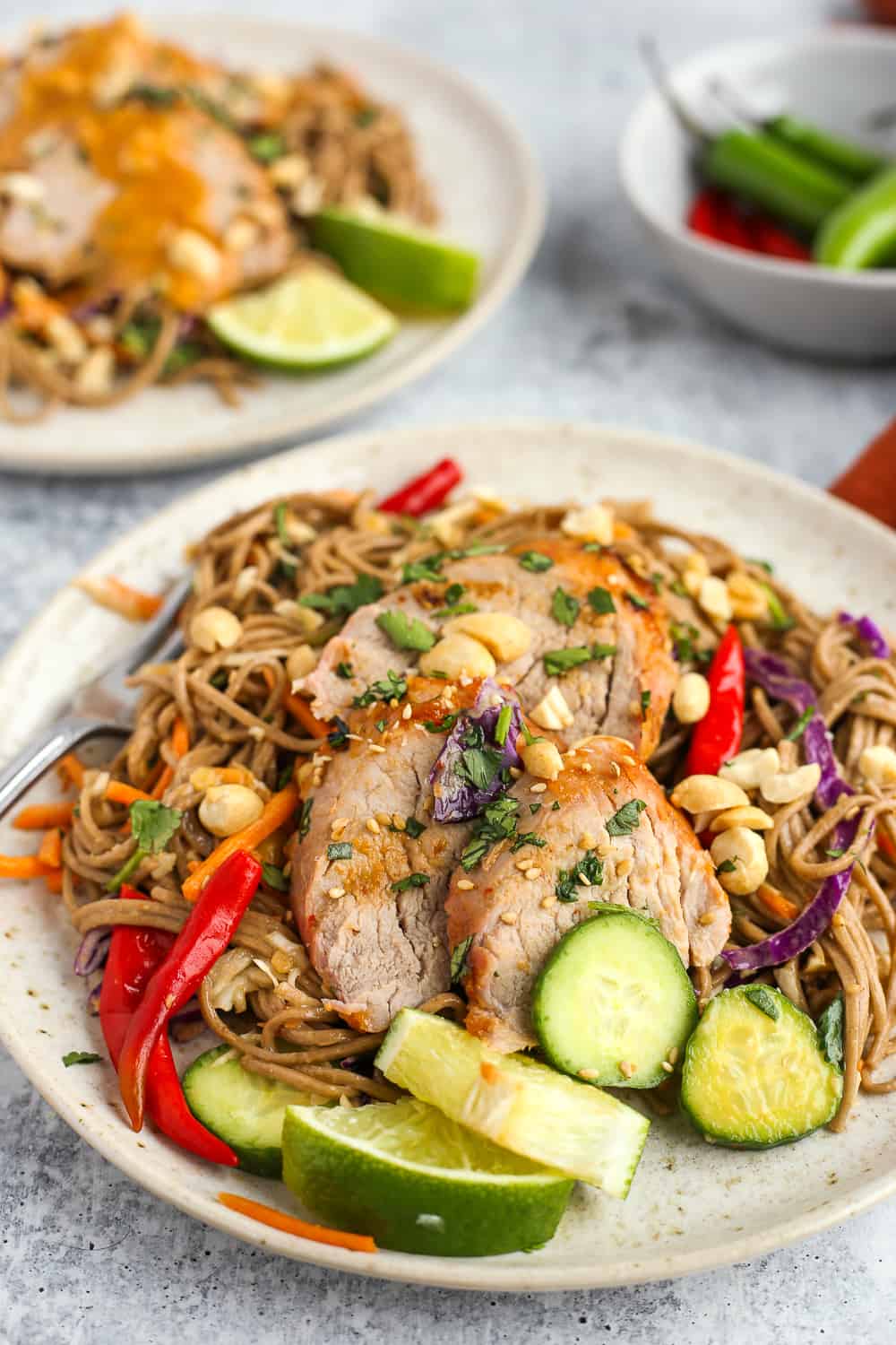 A plated dish of chilled soba noodles with peanut sauce and pork, garnished with lime wedges and red Thai chili peppers