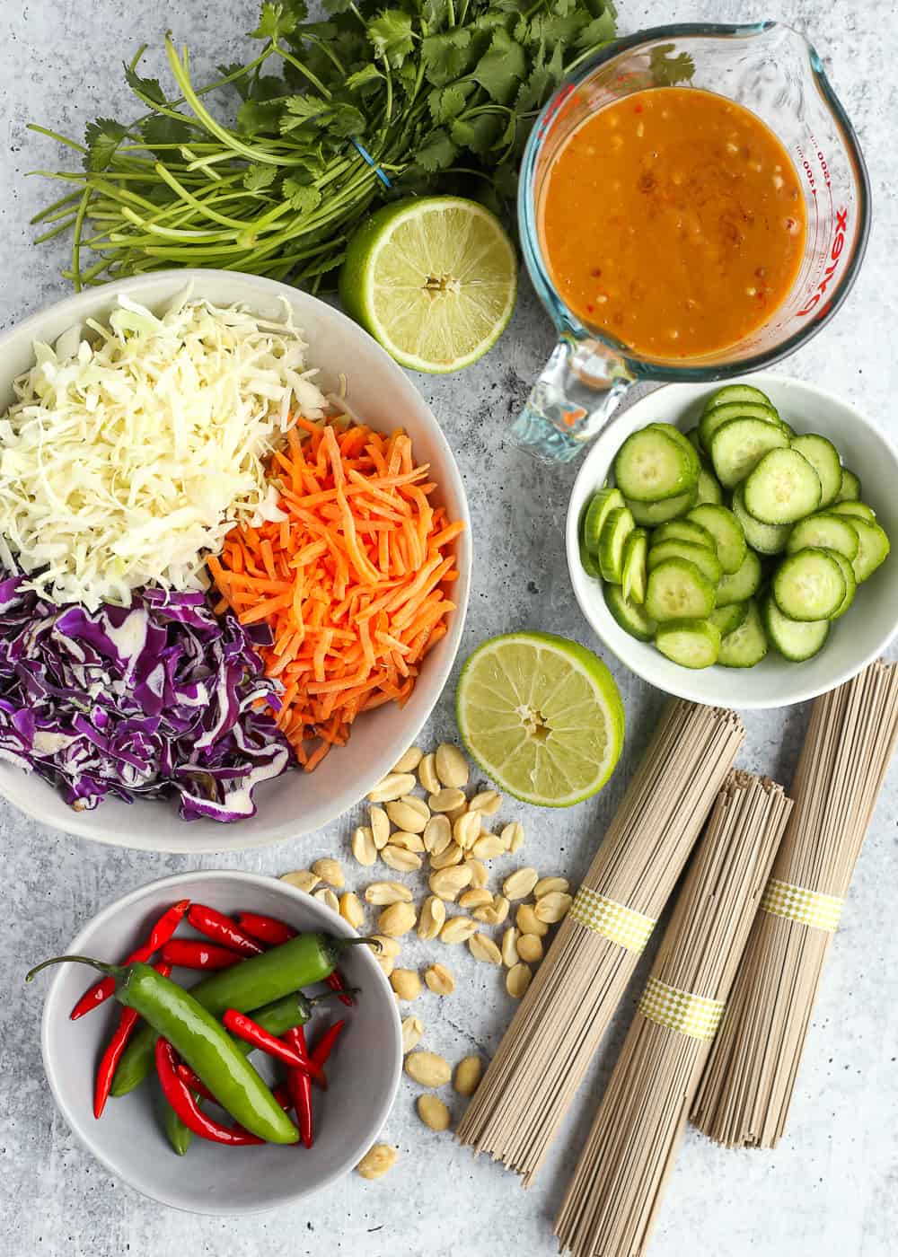 An overhead view of shredded cabbage and carrots in a large bowl, sliced cucumbers in a smaller bowl, peanut sauce in a measuring cup, and additional ingredients surrounding the dishes on a light grey countertop