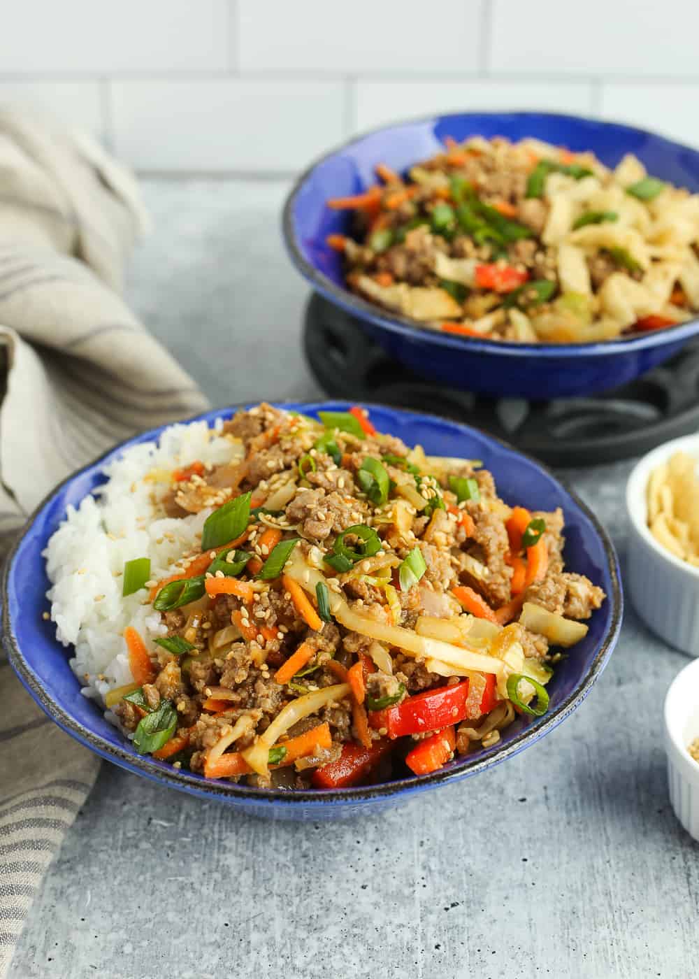 Two blue bowls on a kitchen countertop filled with a ground pork egg roll in a bowl recipe, served with white rice and garnished with sliced green onions and sesame seeds