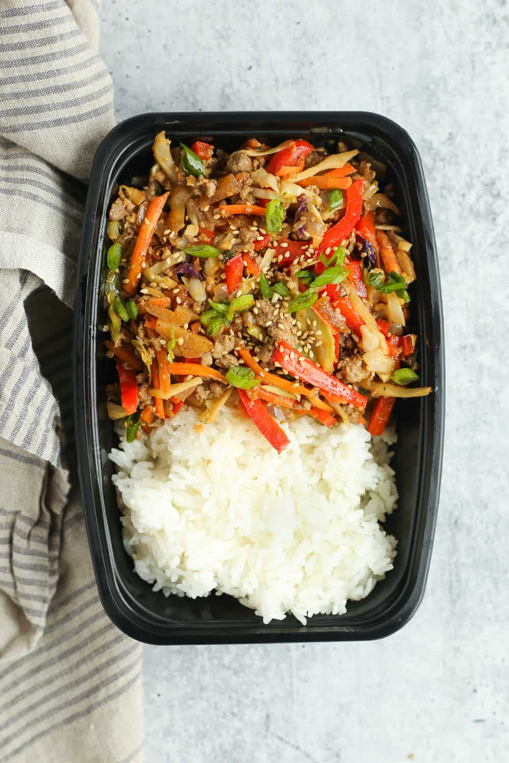 Overhead image of an egg roll in a bowl recipe packed in a black plastic meal prep container with steamed white rice