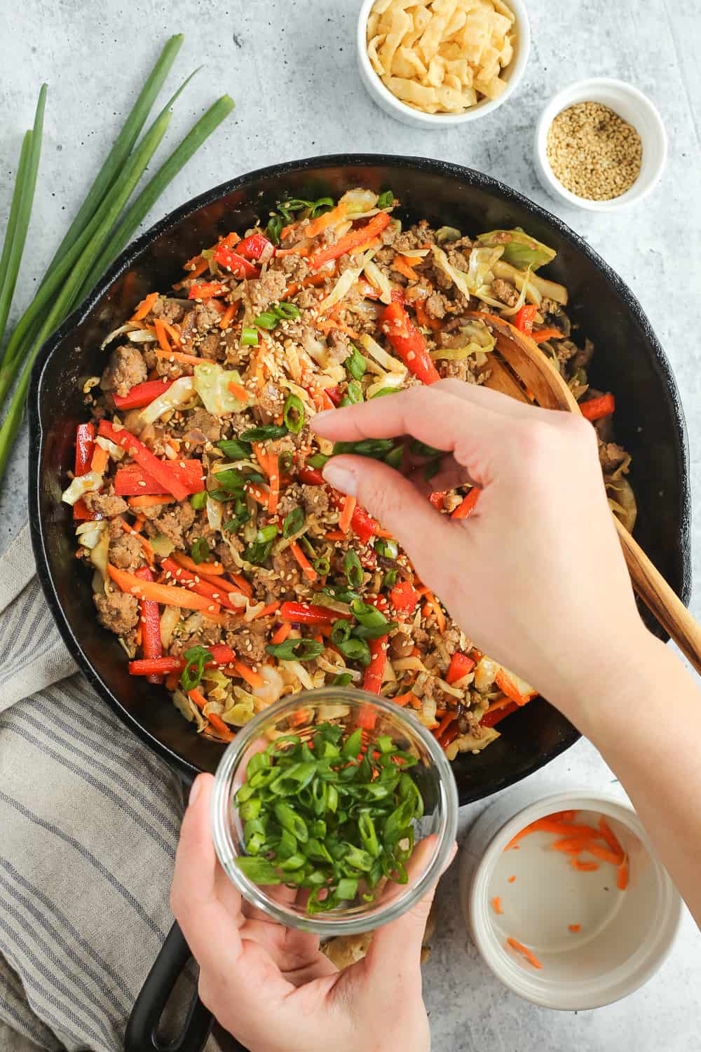 A woman's hand sprinkles sliced green onions over a cast iron skillet filled with cooked ground pork, cabbage, carrots, and red bell peppers